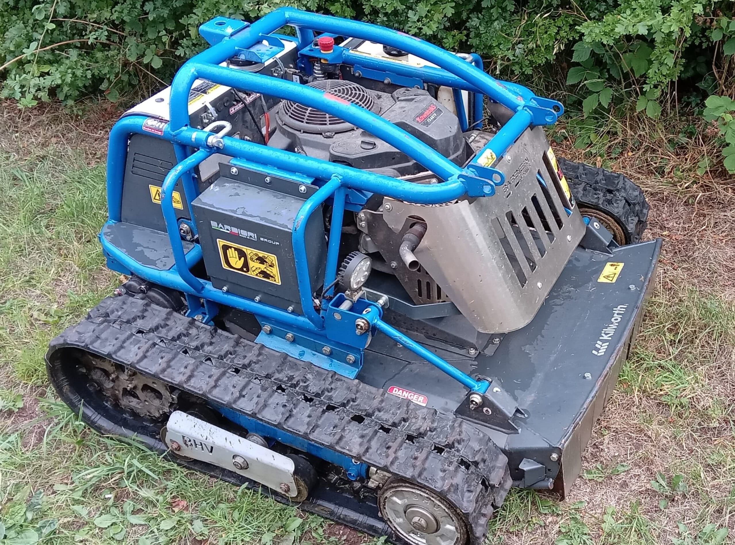 Tracked remote-controlled mower with a flail deck, standing on rough grass beside a hedgerow.