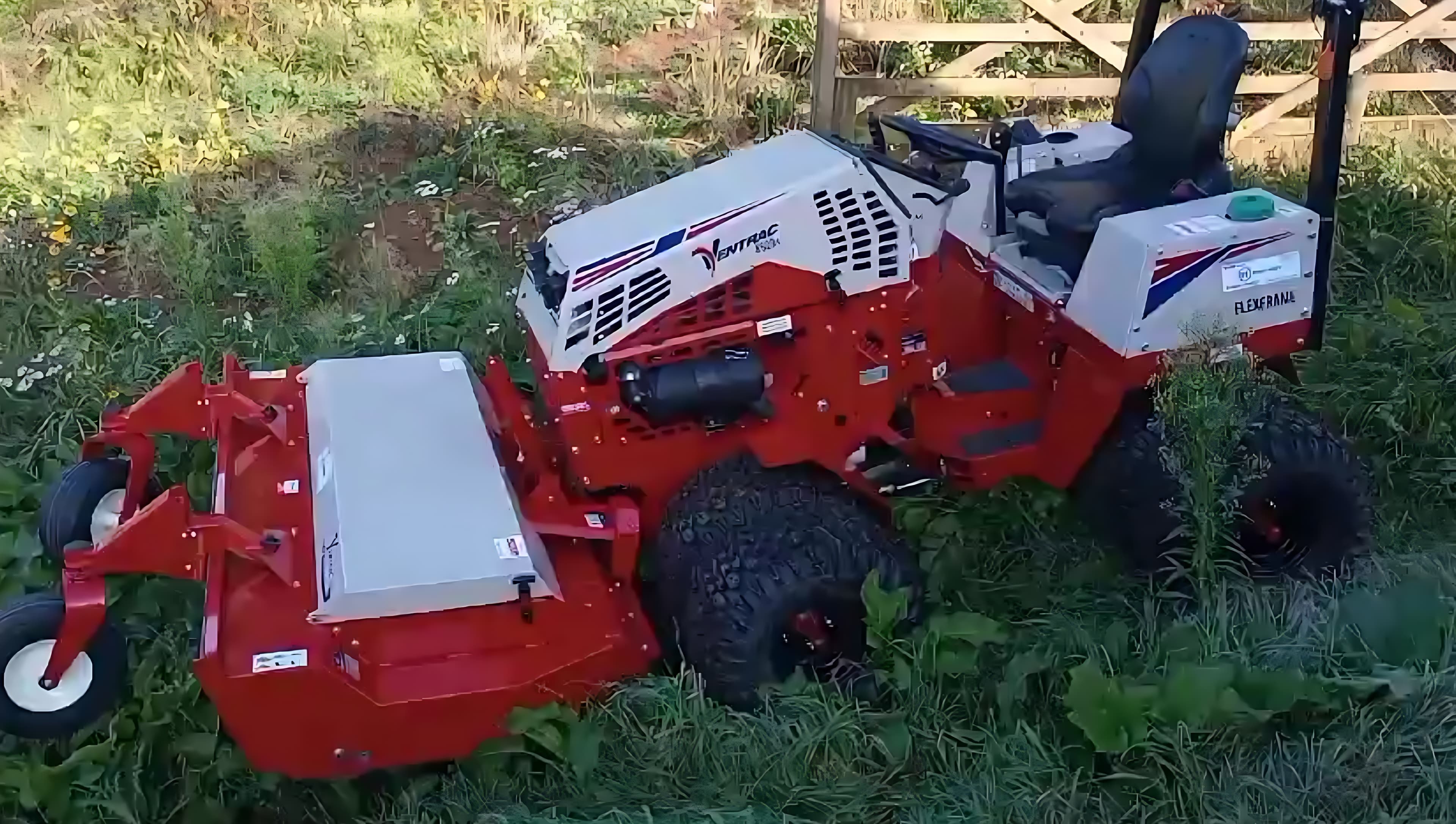 Compact articulated tractor with a rough-cut deck, cutting through dense overgrown vegetation beside a field gate.